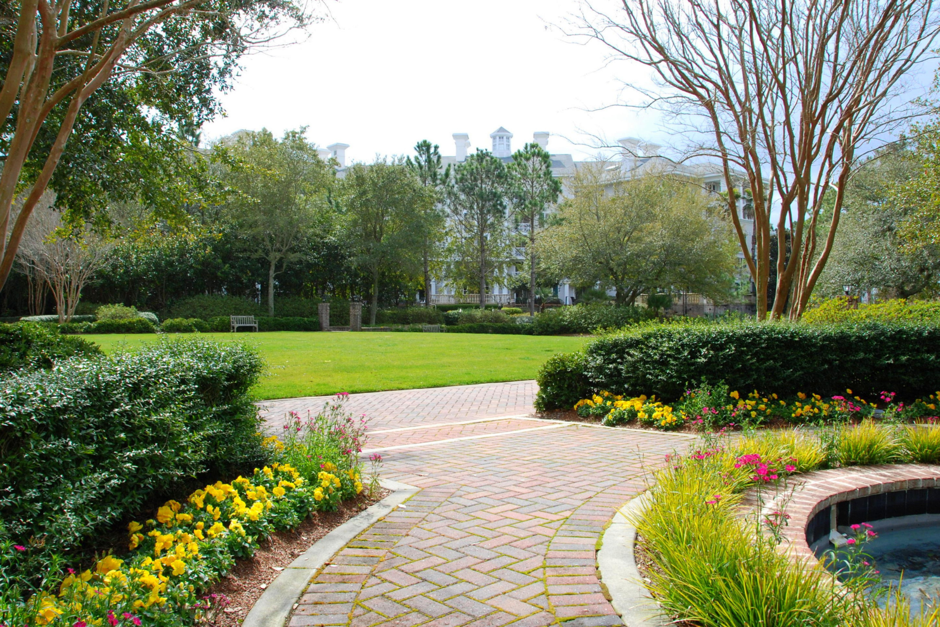 Jardin paysager avec pelouse verte, allée pavée en brique, haies taillées, massifs de fleurs jaunes et roses, grands arbres